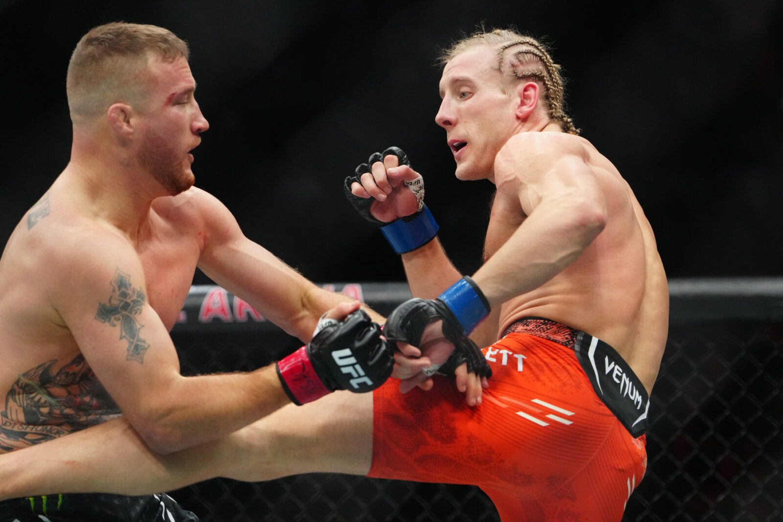 wo mixed martial artists are in the octagon at UFC 324, engaged in an intense match. The fighter on the left is attempting a high kick, while the fighter on the right is in a defensive stance, ready to counter the attack. The audience can be seen in the background, watching the action under the bright arena lights.