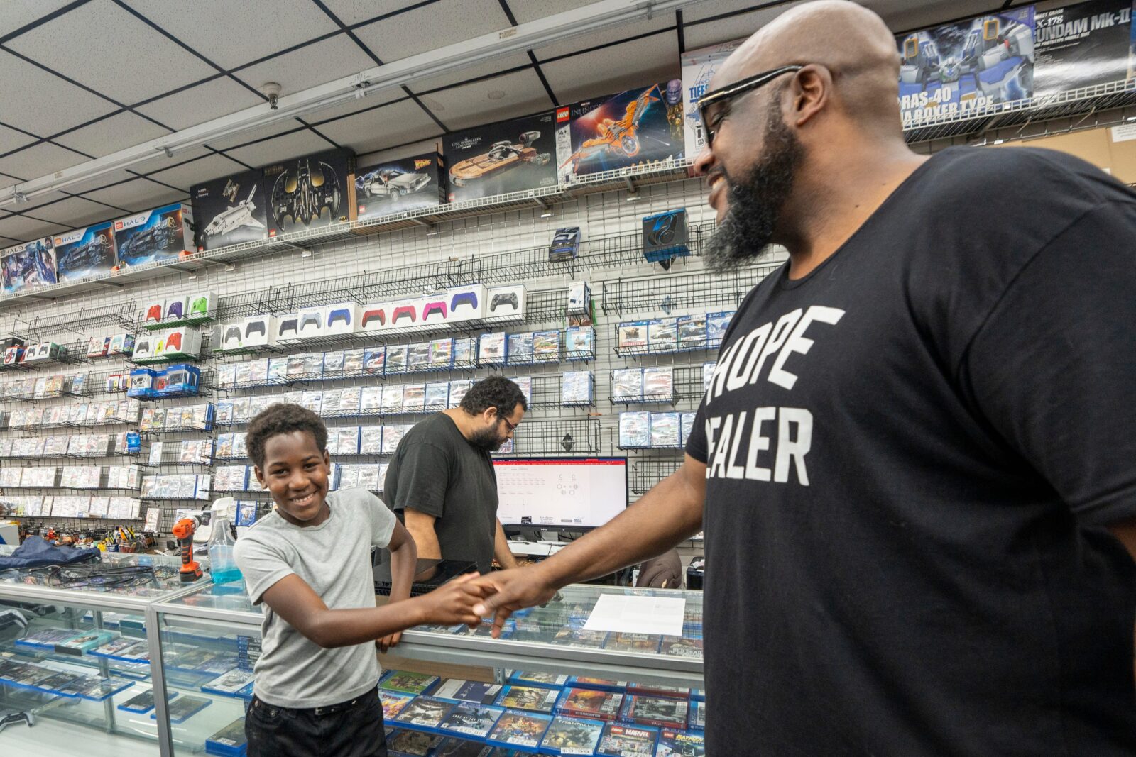 Jul 12, 2024; Paterson, NJ, USA; Shamar Boone (left) shares a laugh with his mentor Jamal Hall (right), at Game Mania as Jonathan Bracerzo (center) helps fix Boone's playstation. Hall became a friend and mentor to Shamar upon his request, while Shamar's father battles a serious illness.